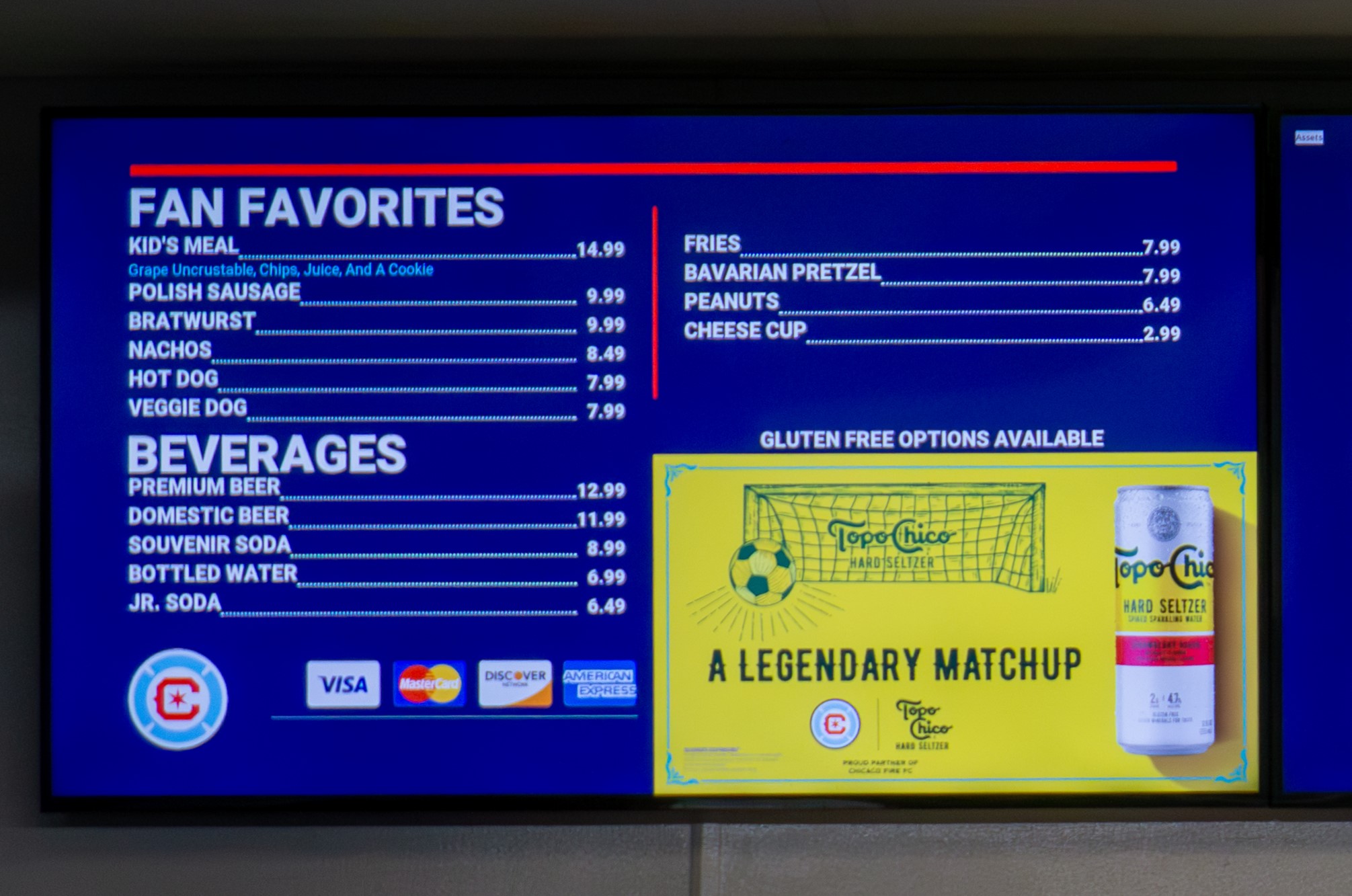 Digital menu signboard at Soldier Field showing a menu. "Gluten free options available" is at the bottom right, above an ad 